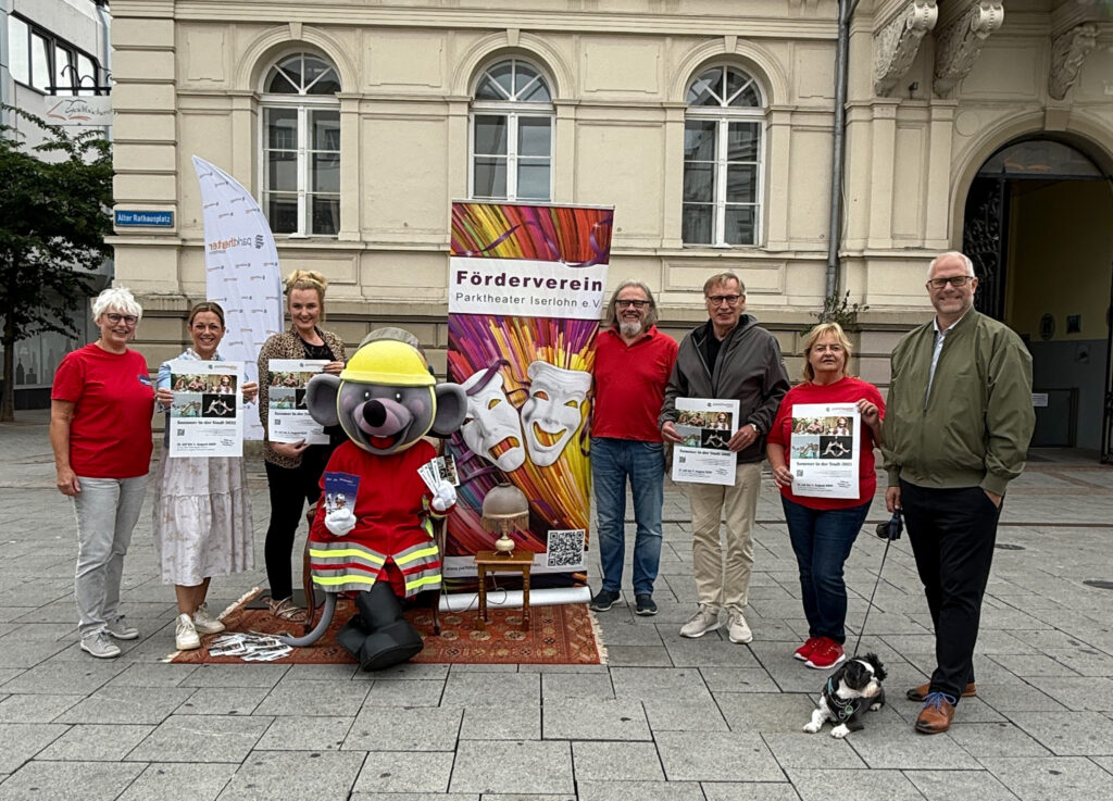 Die Veranstalter stehen mit dem Maskottchen vor dem Alten Rathaus in Iserlohn und präsentieren das Plakat der Veranstaltungsreihe.