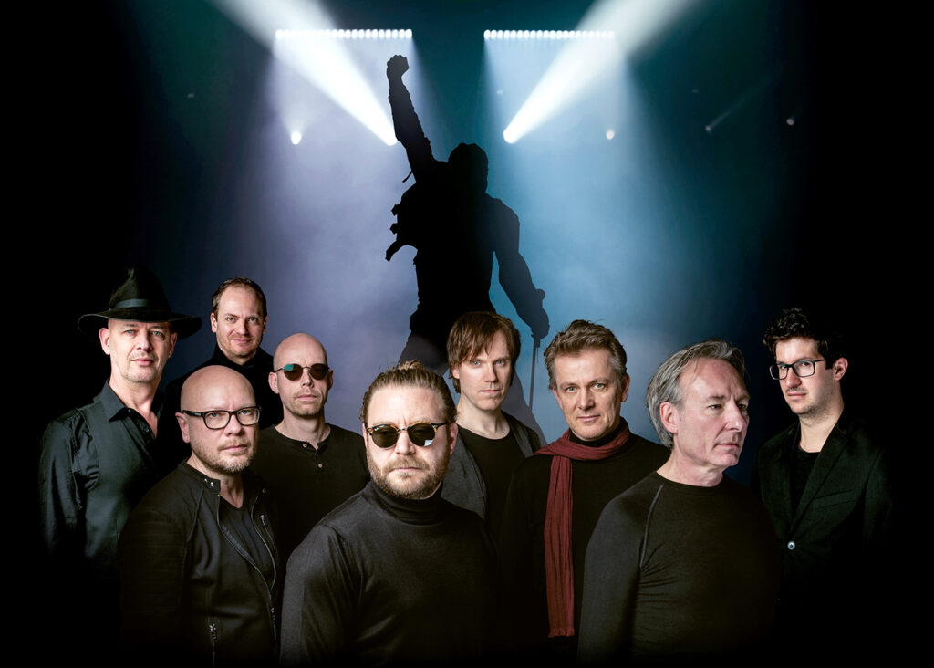 Auf dem Foto sieht man im Vordergrund die Band und im Hintergrund einen Mann, der seinen Arm in die Luft streckt.