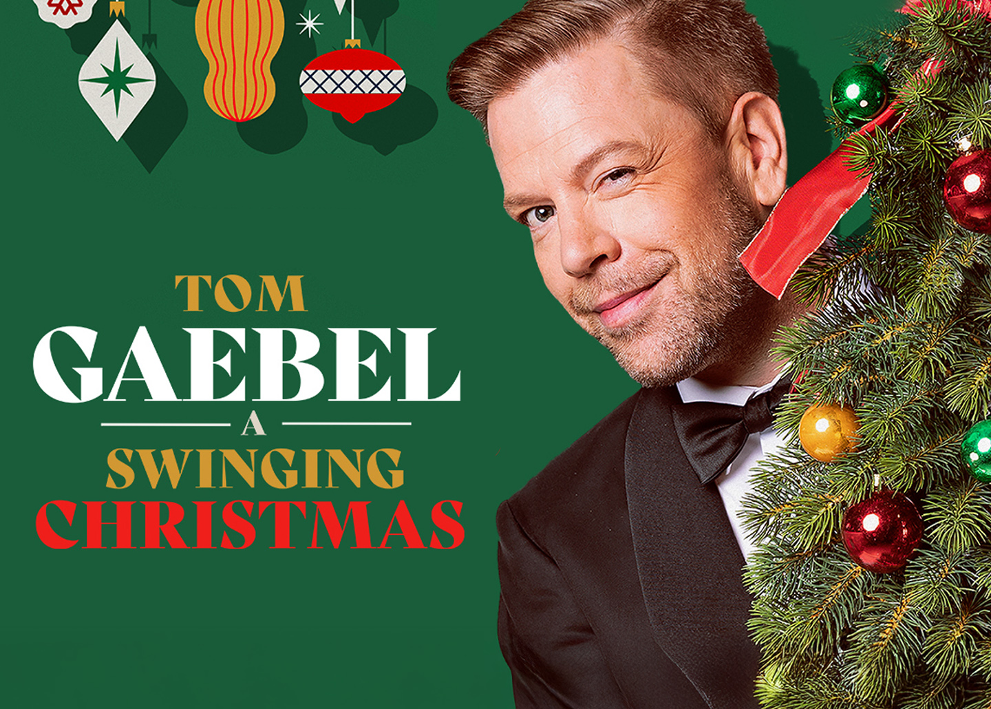 Tom Gaebel hat einen geschmückten Tannenbaum im Arm und zwinkert schelmisch in die Kamera.