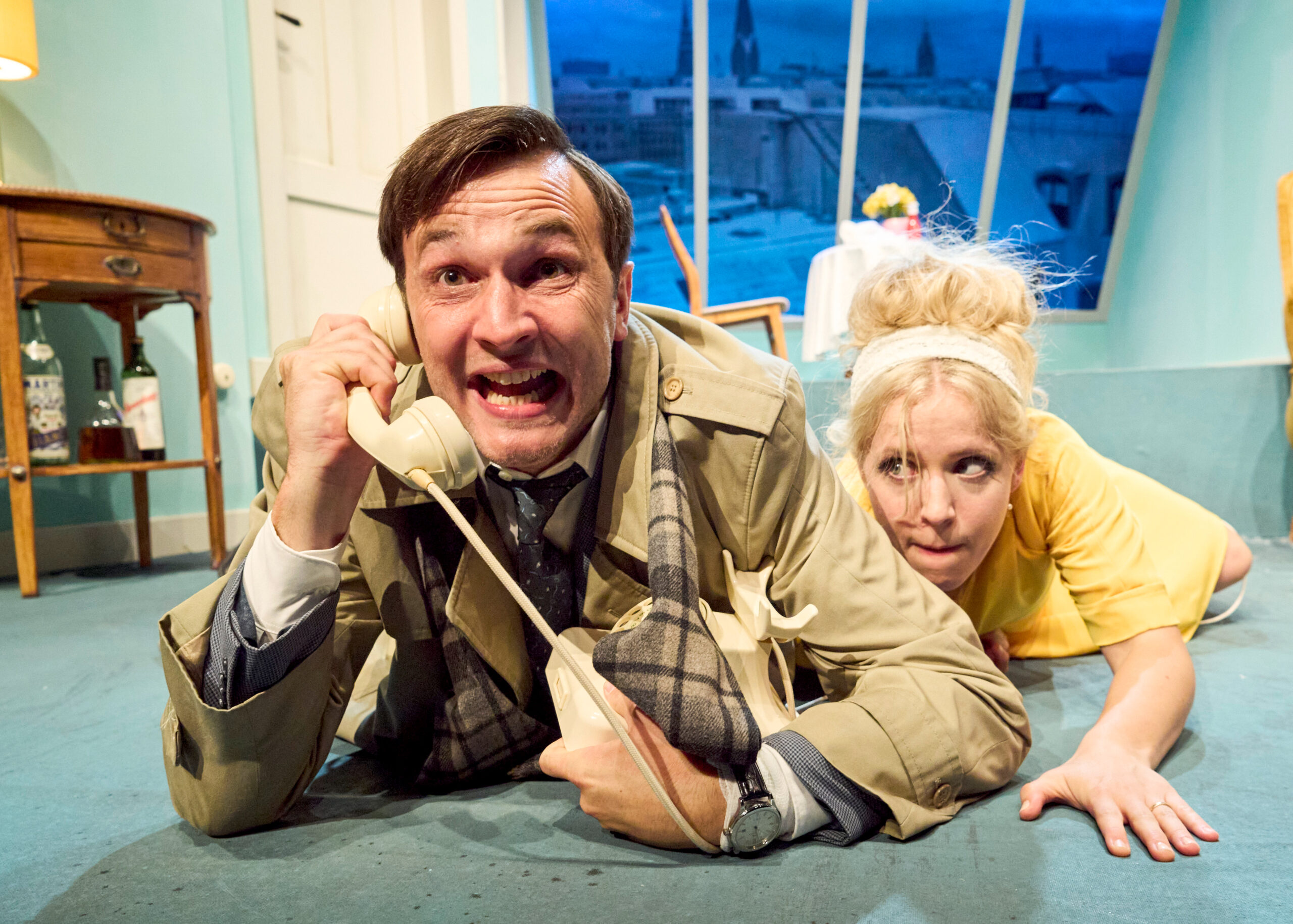 Ein Mann liegt auf dem Boden und telefoniert mit einem Kabel-Telefon. Hinter ihm schleicht sich eine Frau an.