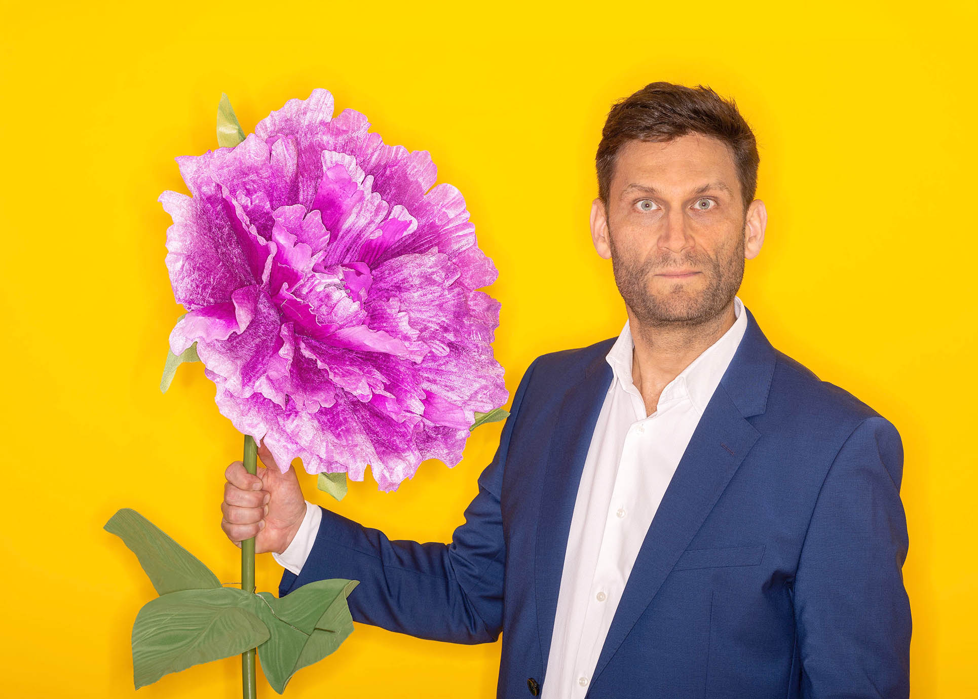 Christian Ehring lächelt in die Kamera und hält dabei eine überdimensional große Kunstblume in der Hand