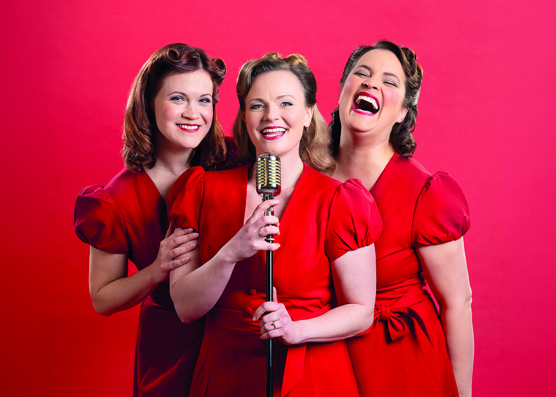 Drei in Rot gekleidete Damen stehen hinter einem Mikrofon vor einem ebenfalls roten Hintergrund