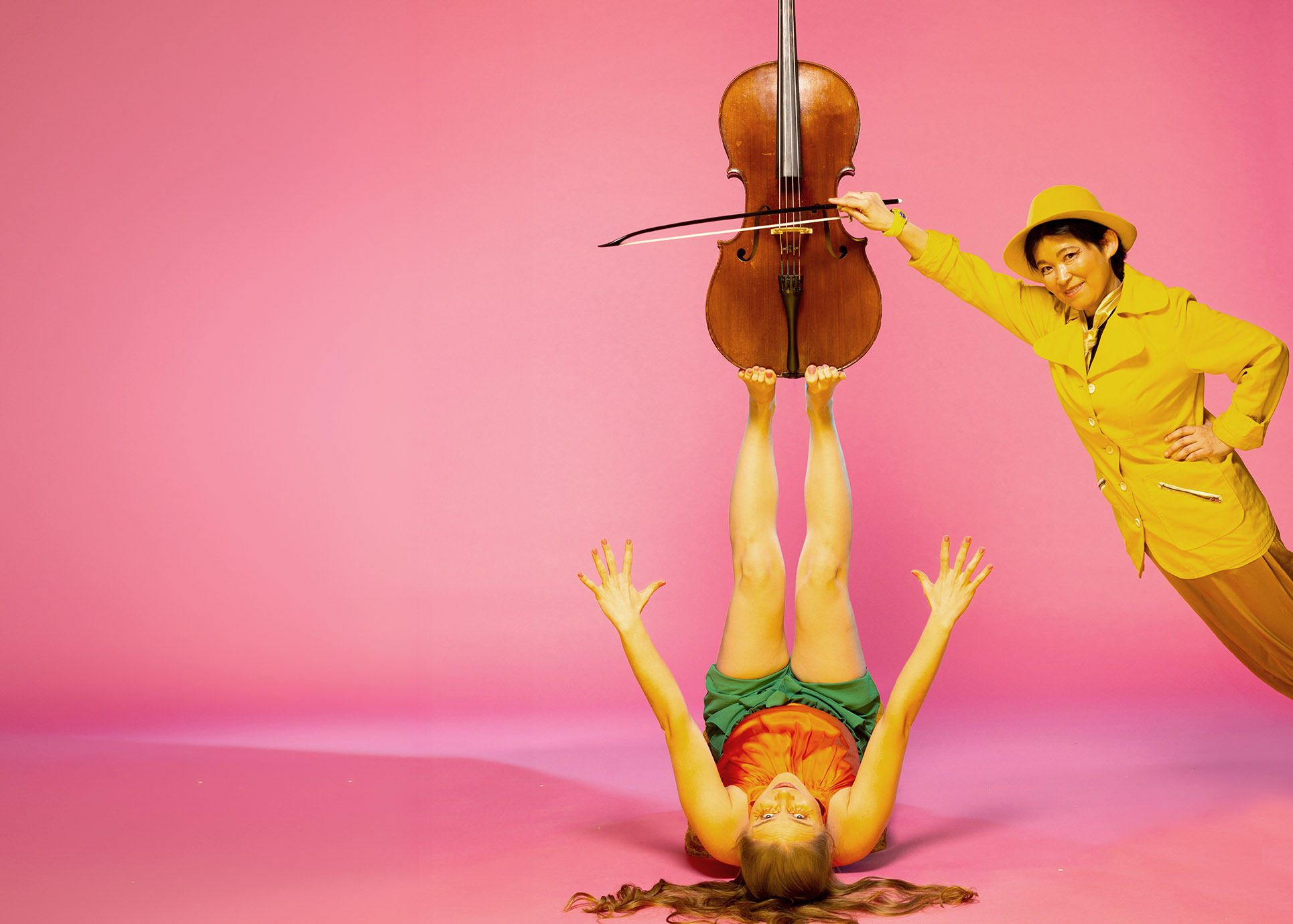 Auf dem Bild liegt eine Dame auf dem Rücken und balanciert eine Violine auf Ihre Füßen. Eine weitere Dame, ganz in in gelb gekleidet, lehnt sich an diese an.