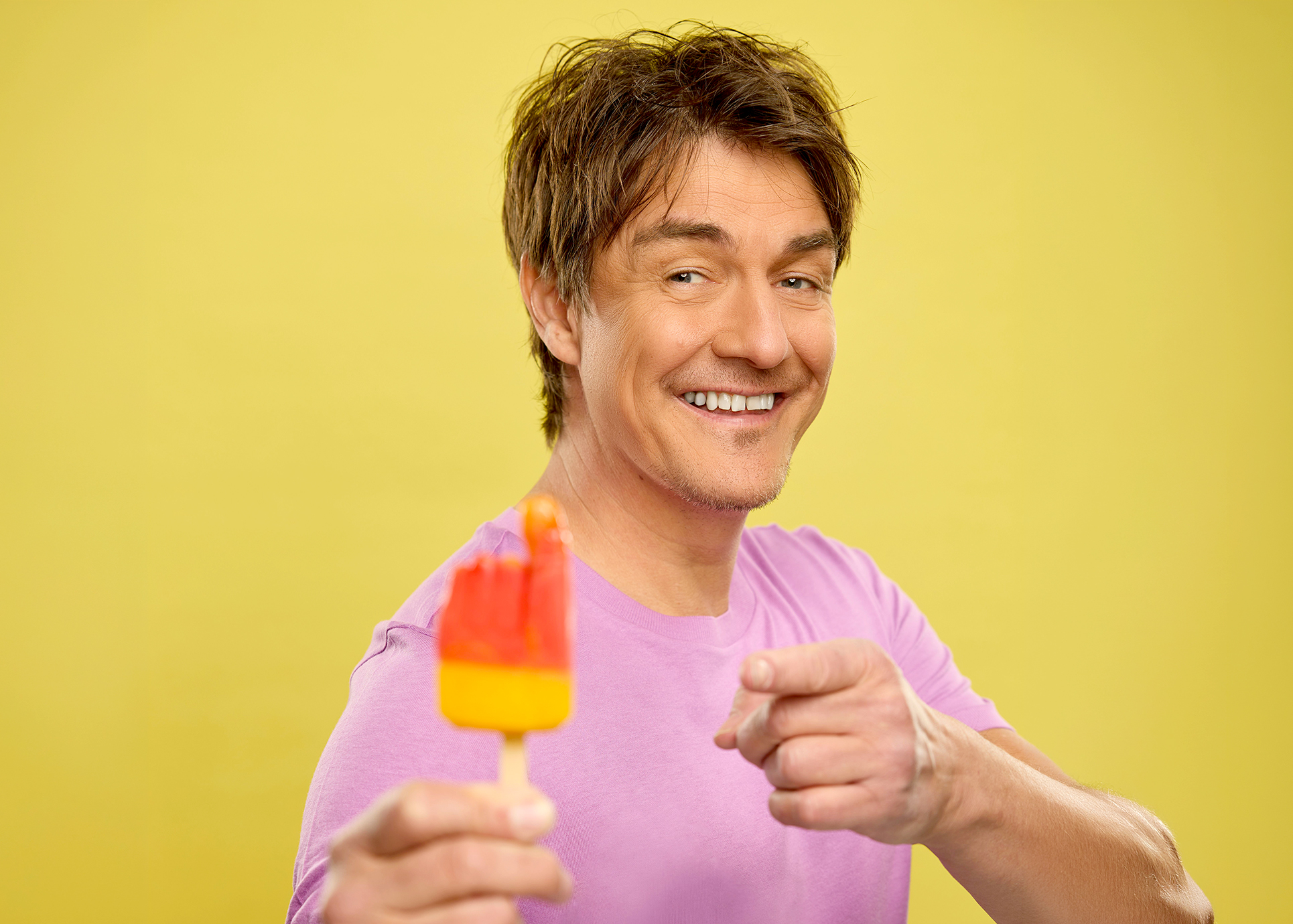 Matze Knop hält ein "Flutschfinger"- Eis in seiner Hand und lächelt in di Kamera