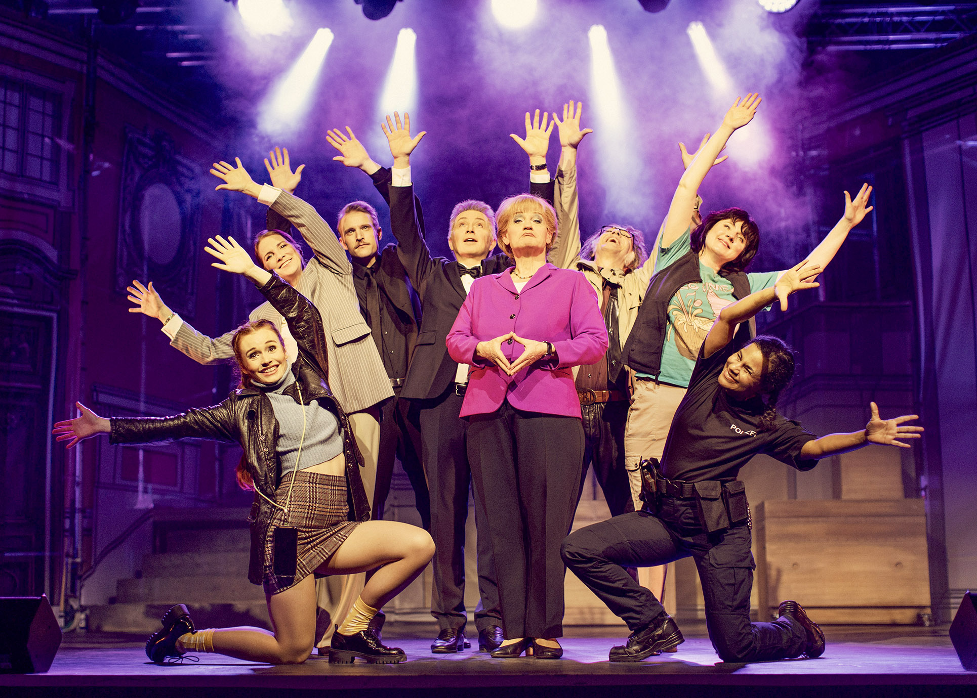 Miss Merkel steht in der Mitte der Bühne umgeben von mehreren Darstellern und deutet mit den Händen ihre bekannte Rauten- Pose an.