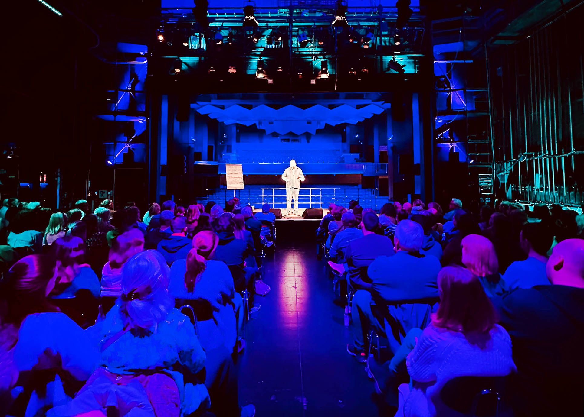 Bunt beleuchtete Hinterbühne im Industriecharme eines Theater, seitlich die Bühne mit einem Poetry-Slammer, davor volle Publikumsreihen.