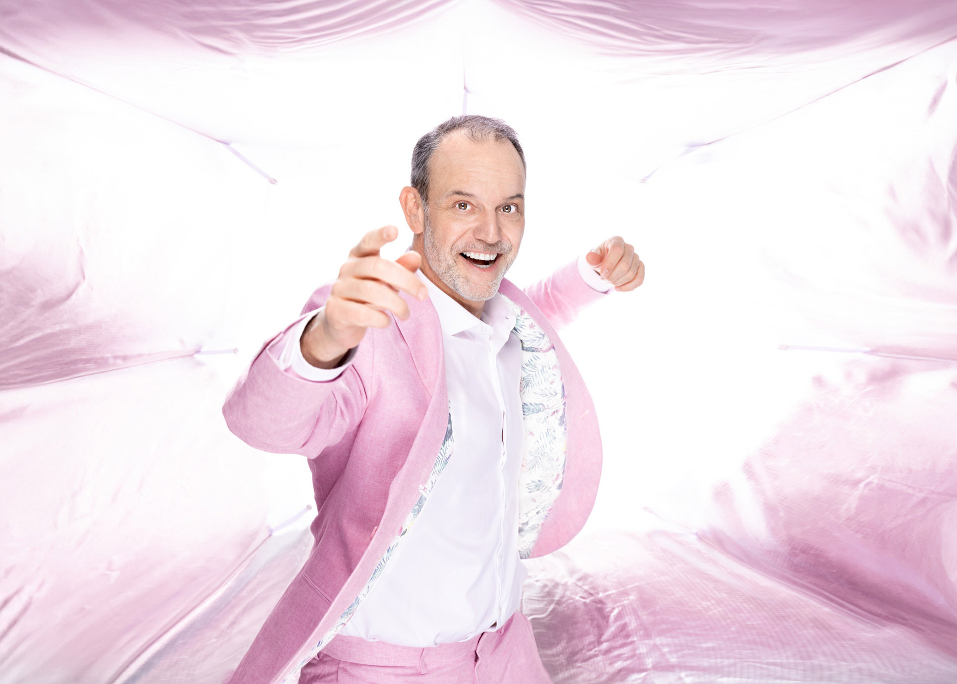 René Steinberg zeigt lachend in die Kamera. Er trägt einen rosafarbenen Anzug und steht vor einem kitschigen, ebenfalls rosafarbenen Hintergrund.
