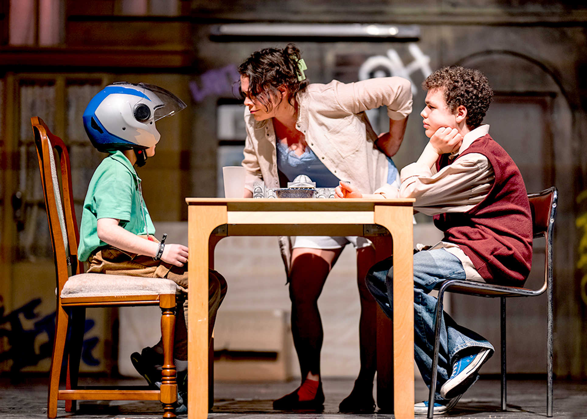 Rico sitzt mit seinem Freund am Tisch, eine Frau beugt sich mit einem strengen Gesichtsausdruck über ihn.