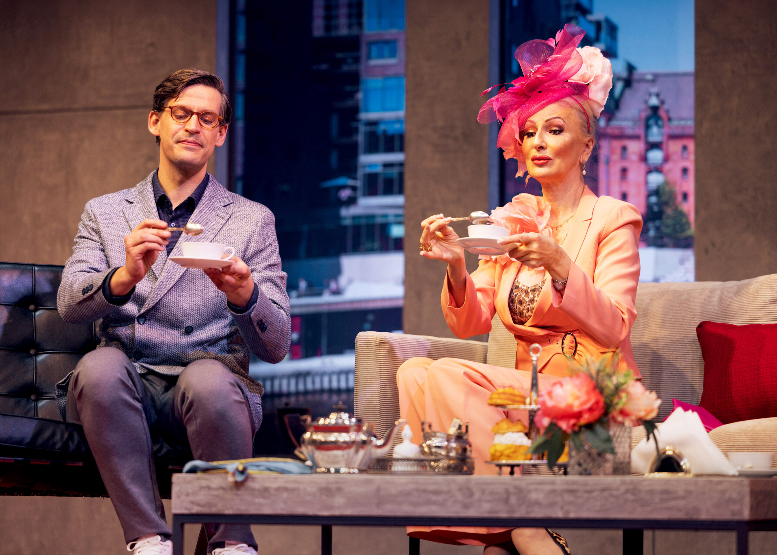 Das Bild zeigt einen einen Mann und eine auffällig gekleidete Frau mit Hut im Wohnzimmer während des Teetrinkens.