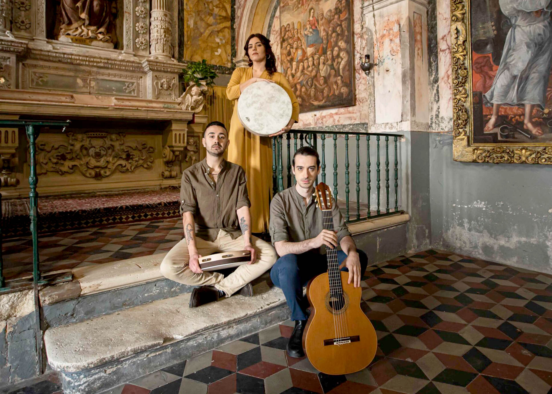 Auf dem Bild sind 23Musiker zu sehen die ihre Instrumente in der Hand halten. Sie stehen dabei in einer historischen Räumlichkeit, die an eine Kirche oder einen Ballsaal erinnern könnte