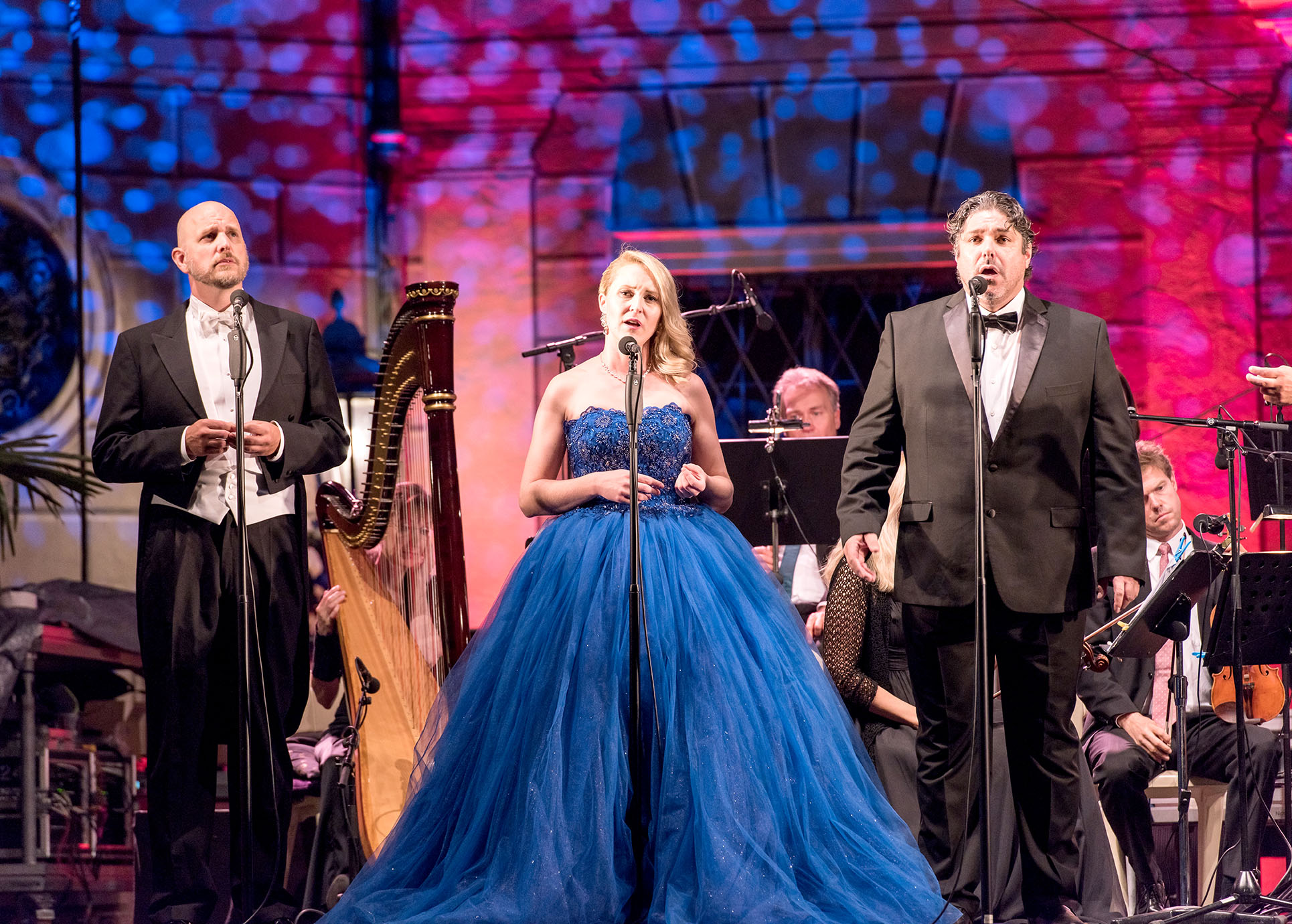 Auf der Bühne steht eine Opernsängerin in einem blauen Ballkleid in der Mitte. Neben ihr stehen zwei Opernsänger. Im Hintergrund sieht man einen Teil des Orchesters.