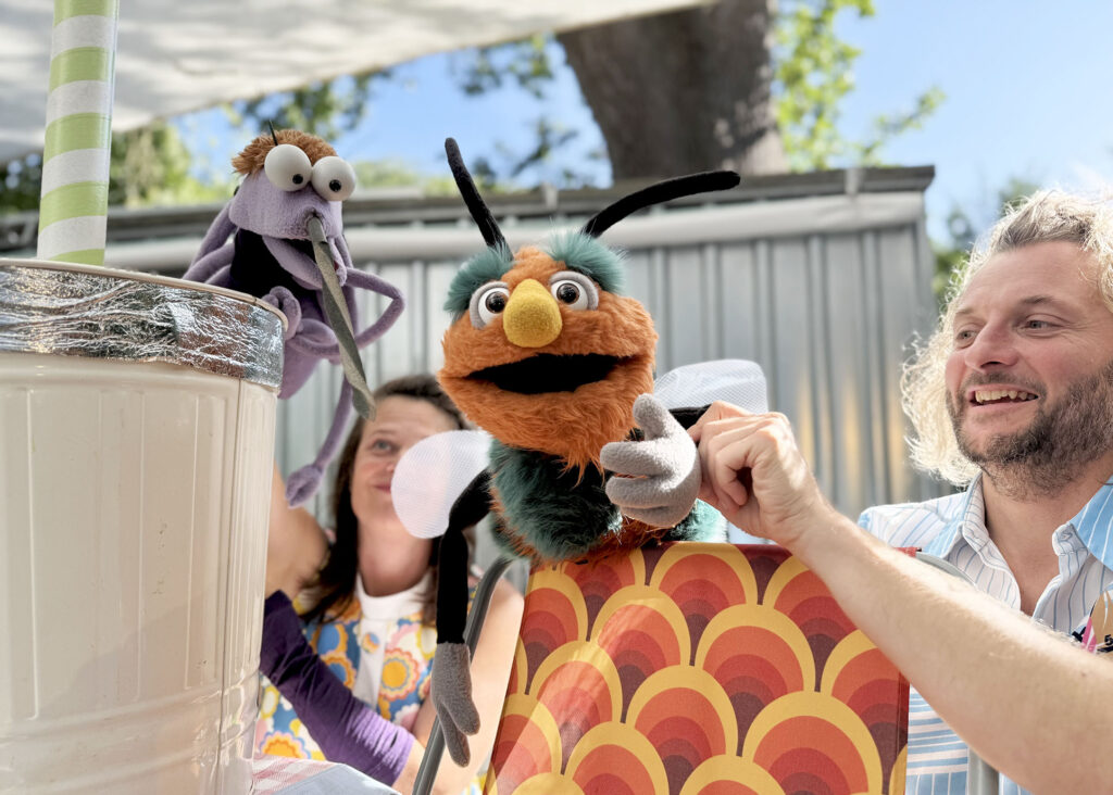 Zwei Handpuppen lächeln in die Kamera, dahinter ist versteckt der Puppenspieler zu sehen.