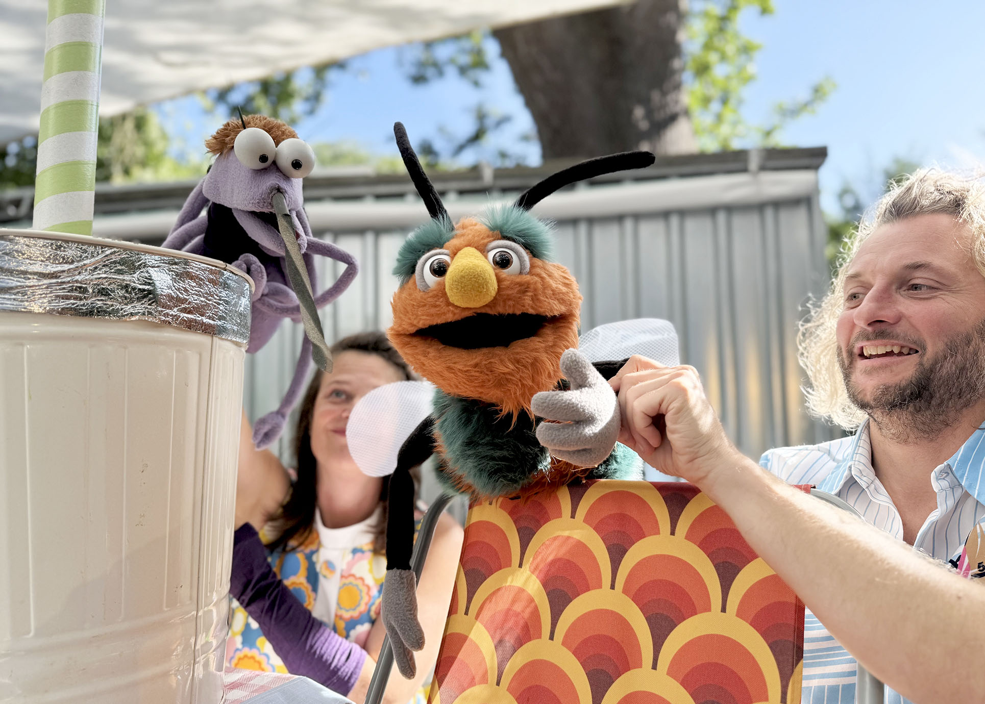 Zwei Handpuppen lächeln in die Kamera, dahinter ist versteckt der Puppenspieler zu sehen.